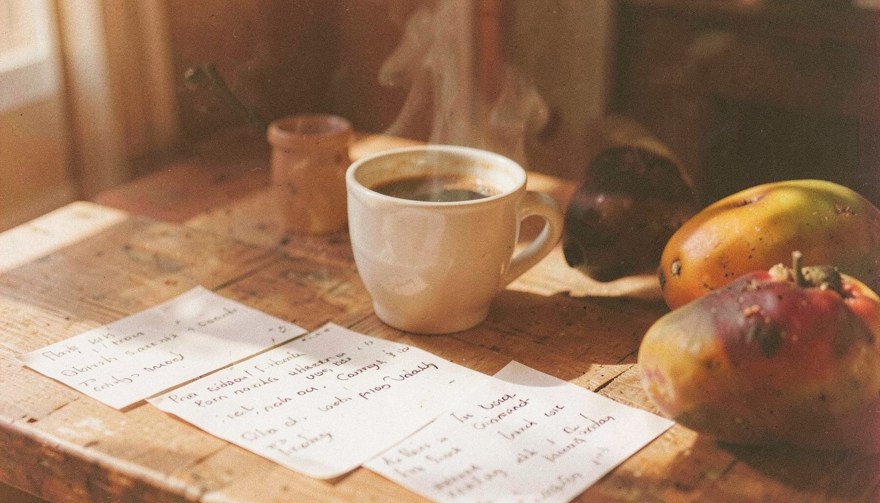Meja kayu yang disinari matahari dengan catatan resep tulisan tangan, secangkir kopi mengepul, dan beberapa buah tropis matang