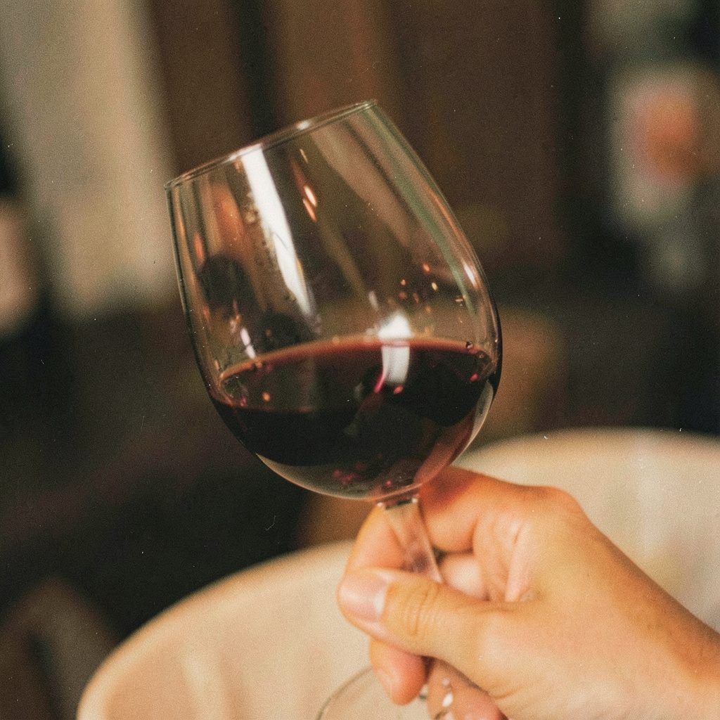 Tangan memutar gelas wine dengan cairan burgundy yang menangkap cahaya, suasana hangat dan akrab